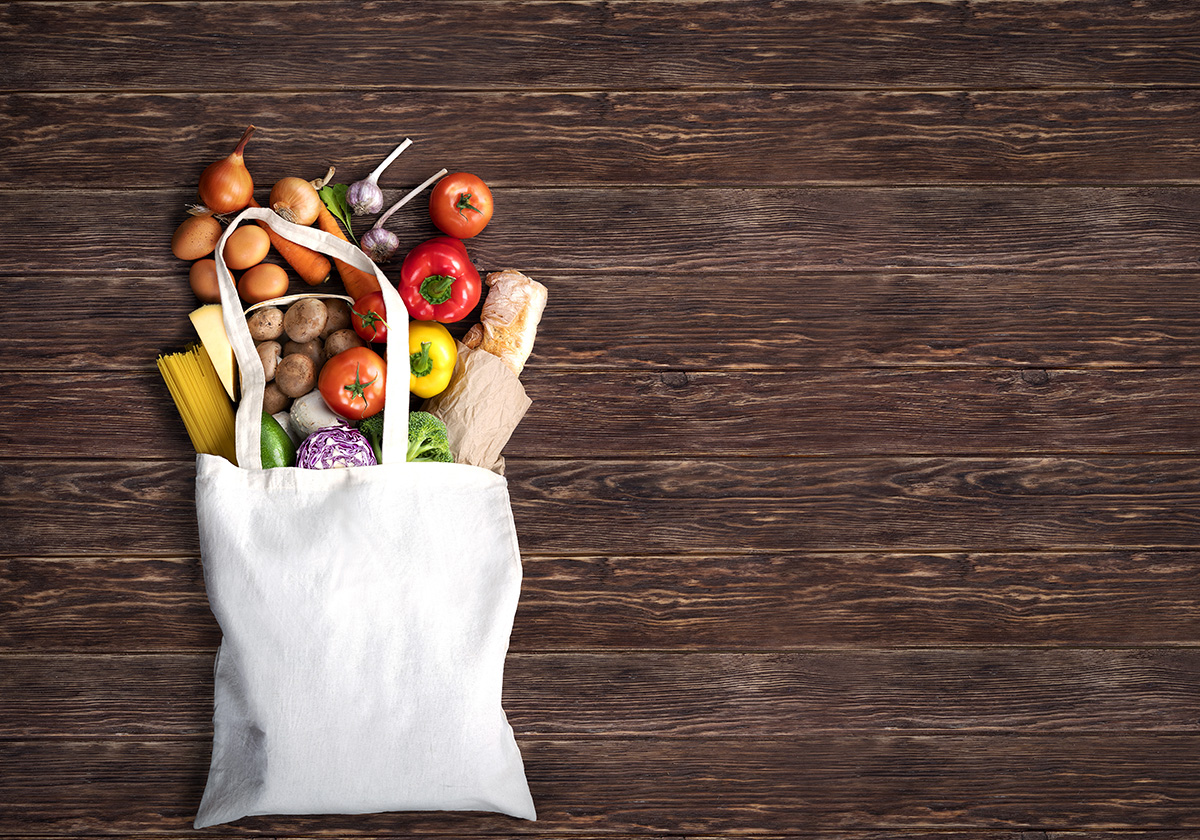 Canvas bag of grocers on a wood panel background with fruits, vegetables and breads coming out of the top of the bag.